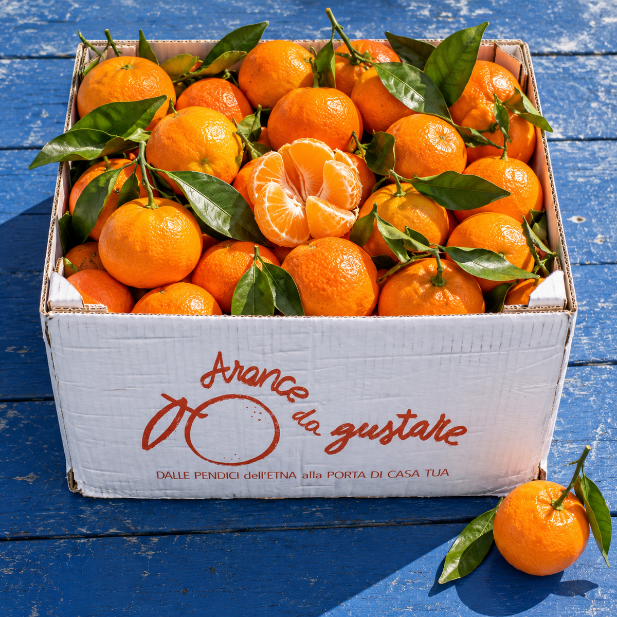 Cassetta Arancedagustare piena di clementine e mandarini con foglie, su un tavolo di legno blu al sole